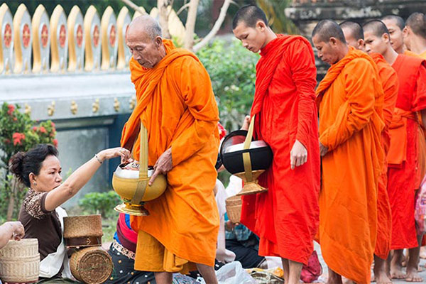 A group of Buddhist monks in robes walk in a line holding urns in an receiving food.